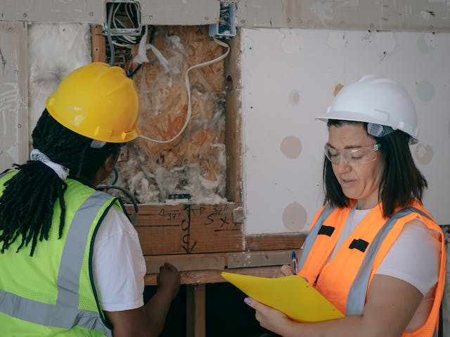 two-people-in-hard-hats-and-safety-vests-inspecting-the-wiring-and-insulation-of-a-wall